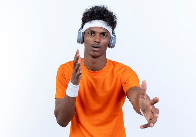 Hombre deportivo afroamericano joven confundido con diadema y muñequera y auriculares extendiendo la mano a la cámara aislada sobre fondo blanco