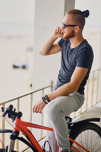 El hombre deportivo barbudo se sienta en una bicicleta de montaña roja y sostiene un teléfono inteligente.