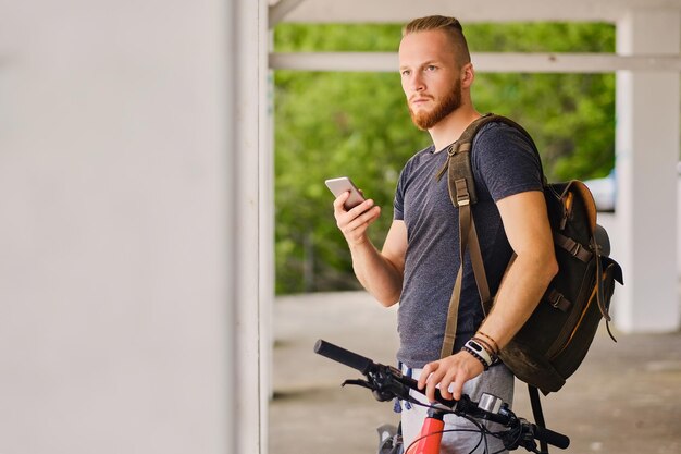 El hombre deportivo barbudo se sienta en una bicicleta de montaña roja y sostiene un teléfono inteligente.