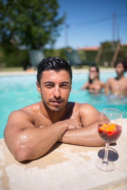 Foto gratuita hombre deportivo bebiendo cócteles brillantes en la piscina. hombre con cabello oscuro y vaso con bebida brillante, mirando a cámara. ocio, amistad, concepto de fiesta