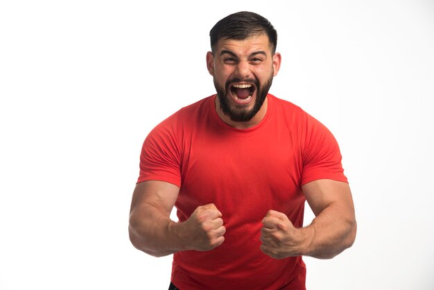 Hombre deportivo en camisa roja demostrando sus músculos del brazo y gritando