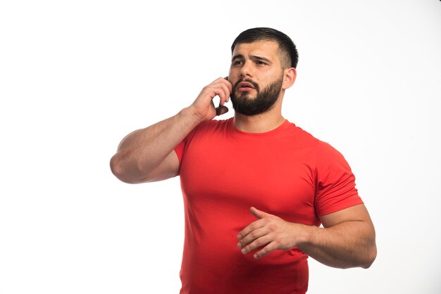 Hombre deportivo en camisa roja hablando con el teléfono y demostrando los músculos de su brazo