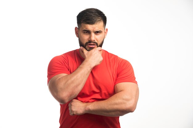 Hombre deportivo con camisa roja que demuestra sus músculos del brazo y parece pensativo.