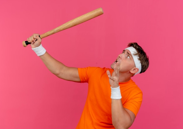 Foto gratuita hombre deportivo guapo joven asustado con diadema y muñequeras sosteniendo un bate de béisbol manteniendo la mano en el aire y mirando hacia arriba aislado en la pared rosa