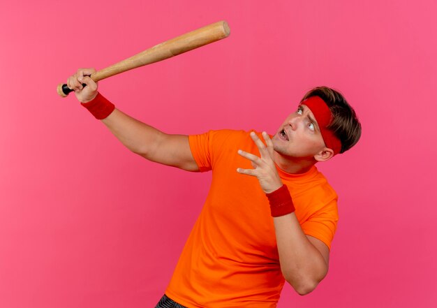 Hombre deportivo guapo joven asustado con diadema y muñequeras sosteniendo un bate de béisbol manteniendo la mano en el aire y mirando hacia arriba aislado en la pared rosa