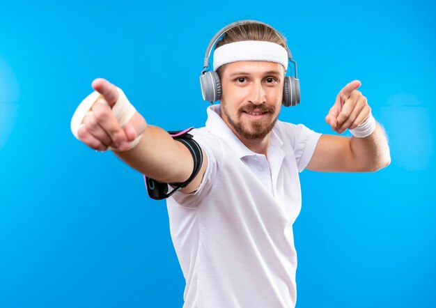 Hombre deportivo guapo joven complacido con diadema y muñequeras y auriculares con brazalete de teléfono apuntando con la muñeca lesionada envuelta con vendaje aislado en el espacio azul