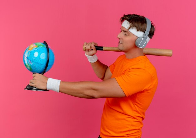 Hombre deportivo guapo joven complacido con diadema, muñequeras y auriculares de pie en la vista de perfil sosteniendo un bate de béisbol en el hombro y el globo y mirando el globo aislado en la pared rosa