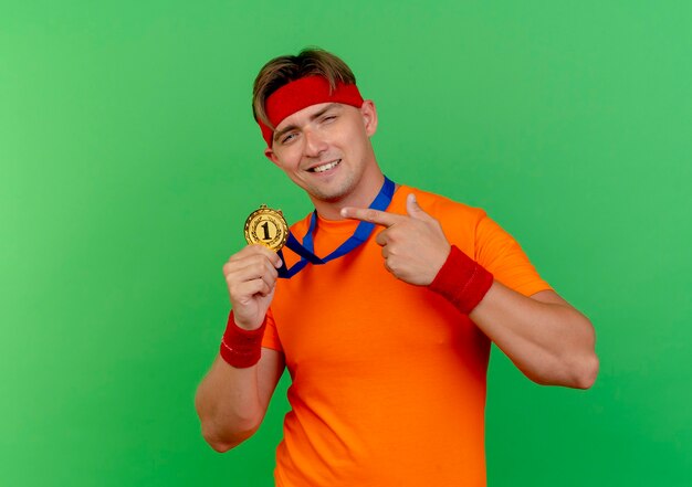 Hombre deportivo guapo joven complacido con diadema y muñequeras con medalla alrededor del cuello sosteniendo y apuntando a la medalla aislada en la pared verde