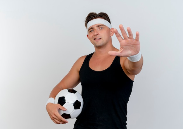 Hombre deportivo guapo joven complacido con diadema y muñequeras sosteniendo un balón de fútbol y estirando la mano hacia el frente aislado en la pared blanca