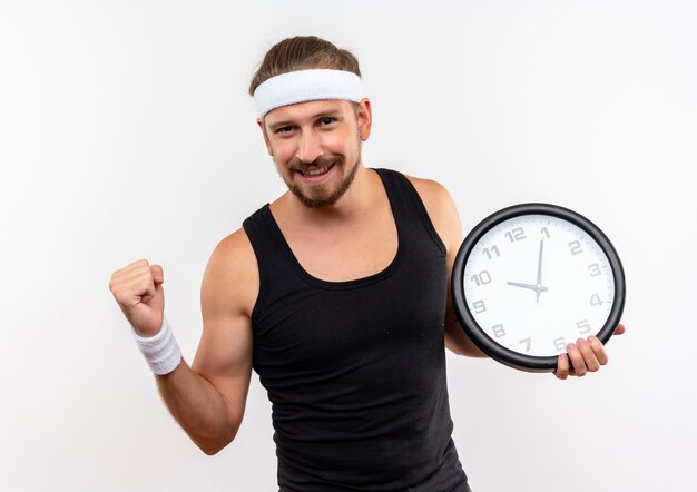 Hombre deportivo guapo joven complacido con diadema y muñequeras sosteniendo el reloj apretando el puño aislado en el espacio en blanco