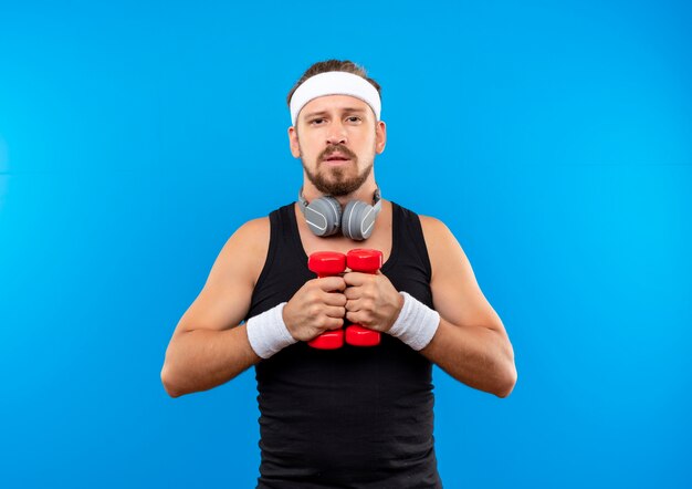 Hombre deportivo guapo joven confiado con diadema y muñequeras y auriculares alrededor del cuello sosteniendo pesas aisladas en la pared azul