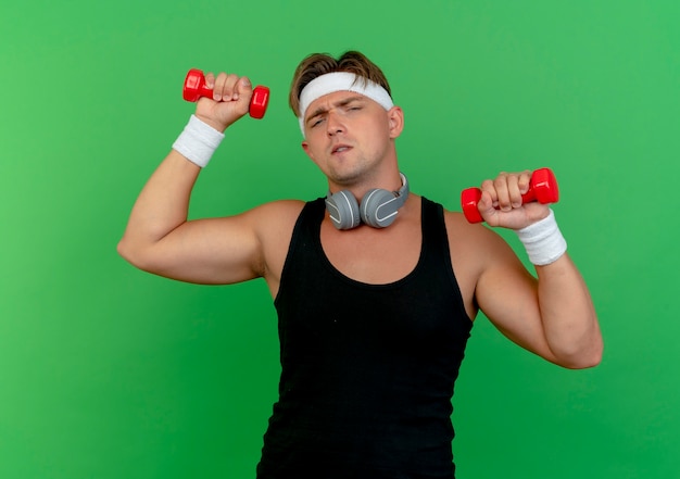 Hombre deportivo guapo joven confiado con diadema y muñequeras con auriculares en el cuello levantando pesas aisladas en verde