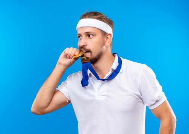 Hombre deportivo guapo joven confiado con diadema y muñequeras y medalla alrededor del cuello sosteniendo y mordiendo la medalla y mirando al lado aislado en la pared azul