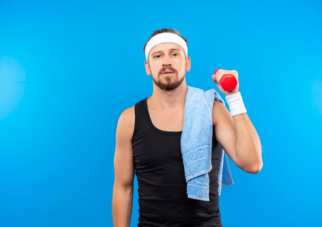 Hombre deportivo guapo joven confiado con diadema y muñequeras sosteniendo mancuernas con una toalla en el hombro aislado en la pared azul con espacio de copia