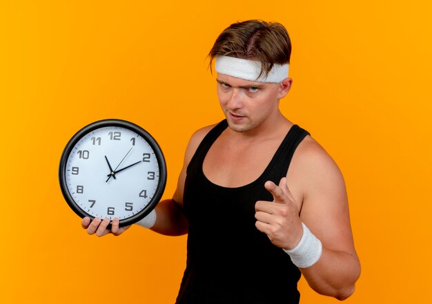 Hombre deportivo guapo joven confiado con diadema y muñequeras sosteniendo el reloj y apuntando aislado en naranja