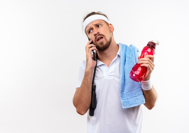 Hombre deportivo guapo joven confundido con diadema y muñequeras hablando por teléfono y sosteniendo una botella de agua con saltar la cuerda y una toalla sobre los hombros aislados en la pared blanca con espacio de copia