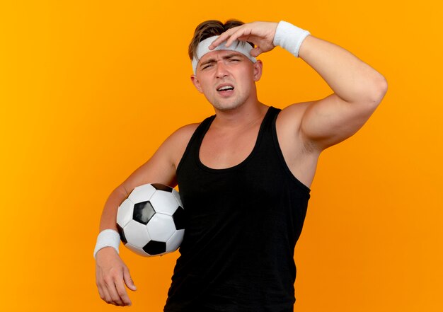 Hombre deportivo guapo joven disgustado con diadema y muñequeras sosteniendo un balón de fútbol y poniendo la mano cerca de la cabeza aislada sobre fondo naranja
