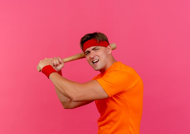 Hombre deportivo guapo joven enojado con diadema y muñequeras con bate de béisbol preparándose para vencer aislado en rosa con espacio de copia