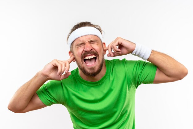 Hombre deportivo guapo joven enojado con diadema y muñequeras poniendo los dedos en los oídos con los ojos cerrados aislados en la pared blanca