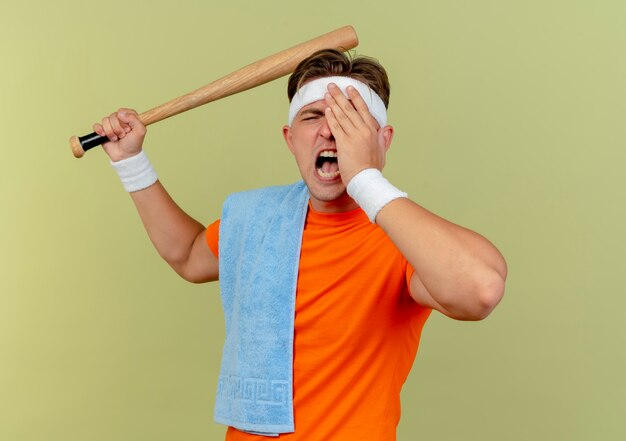 Hombre deportivo guapo joven enojado con diadema y muñequeras con una toalla en el hombro levantando el bate de béisbol preparándose para vencer a alguien poniendo la mano en el ojo aislado en verde oliva