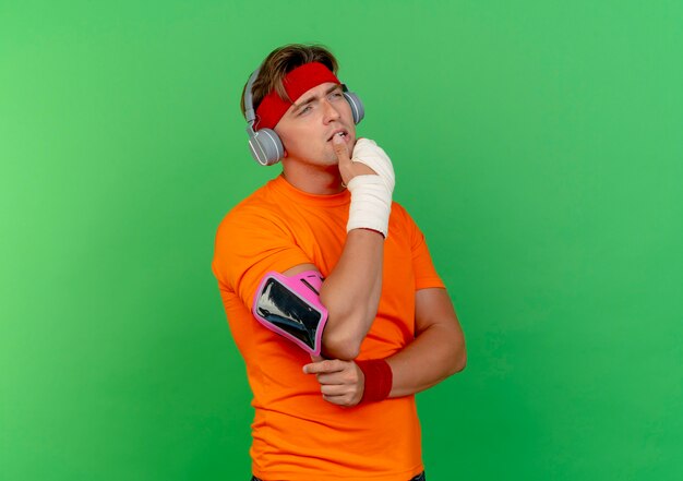 Hombre deportivo guapo joven pensativo con diadema y muñequeras y auriculares y brazalete de teléfono con la muñeca envuelta con una venda poniendo el dedo en el labio mirando directamente