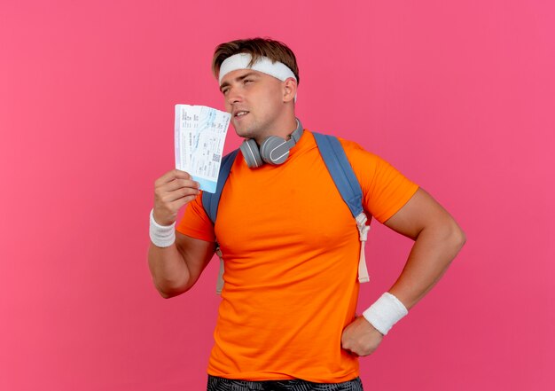 Hombre deportivo guapo joven pensativo con diadema y muñequeras y bolso trasero con auriculares en el cuello sosteniendo boletos de avión mirando de lado con la mano en la cintura