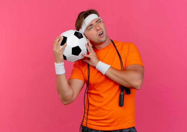 Hombre deportivo guapo joven pensativo con diadema y muñequeras con saltar la cuerda alrededor del cuello sosteniendo un balón de fútbol mirando al lado aislado en la pared rosa