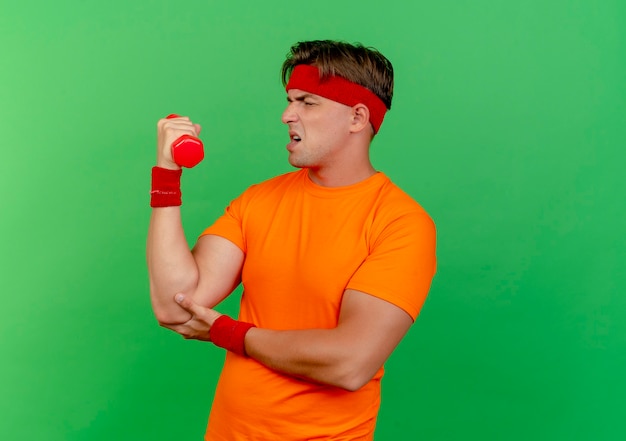 Foto gratuita hombre deportivo guapo joven tenso con diadema y muñequeras levantando mancuernas poniendo la mano debajo del codo aislado en la pared verde