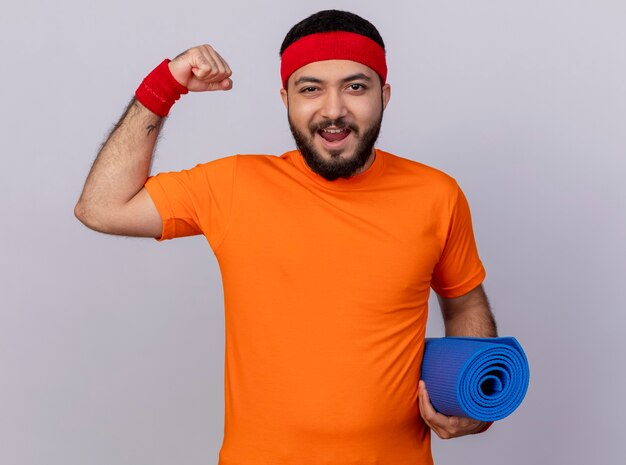 Foto gratuita hombre deportivo joven alegre con diadema y muñequera sosteniendo estera de yoga mostrando un gesto fuerte aislado sobre fondo blanco.