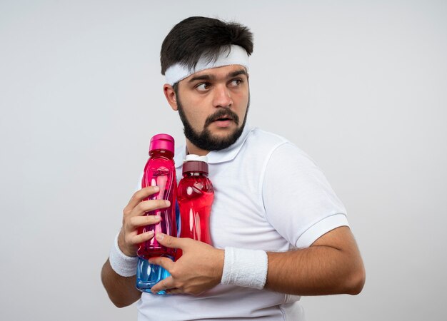 Hombre deportivo joven codicioso con diadema y muñequera sosteniendo botellas de agua aisladas en la pared blanca