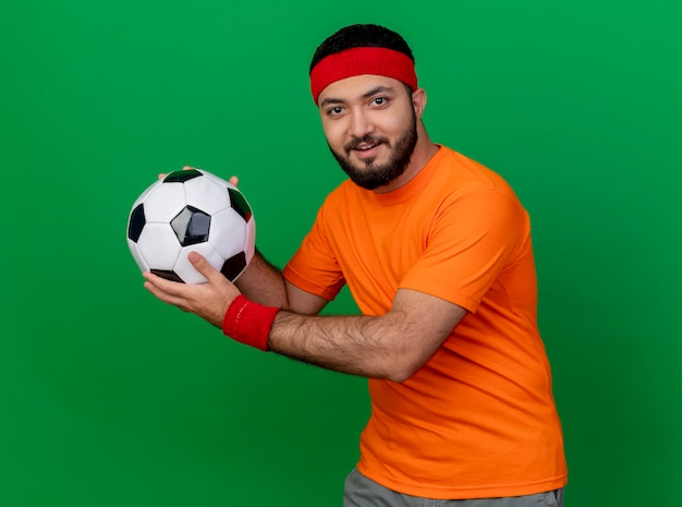 Foto gratuita hombre deportivo joven complacido con diadema y muñequera sosteniendo la bola en el lado aislado sobre fondo verde