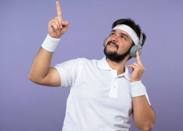 Hombre deportivo joven complacido mirando hacia arriba con diadema y muñequera con auriculares apunta hacia arriba