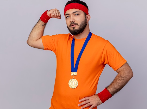 Hombre deportivo joven confiado con diadema y muñequera con medalla poniendo la mano en la cadera mostrando un gesto fuerte aislado sobre fondo blanco.