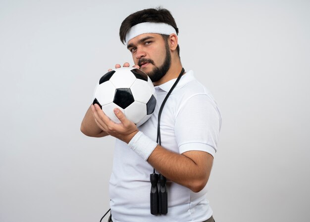 Hombre deportivo joven confiado con diadema y muñequera con saltar la cuerda en el hombro sosteniendo la pelota alrededor de la cara aislada en la pared blanca con espacio de copia