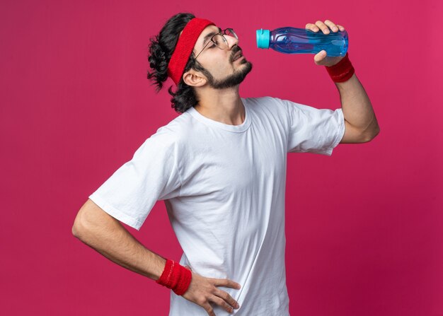 Hombre deportivo joven confiado con diadema con muñequera sosteniendo una botella de agua poniendo la mano en la cadera