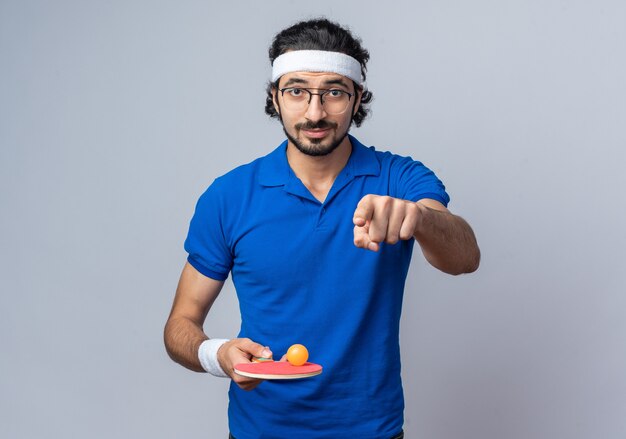 Hombre deportivo joven confiado con diadema con muñequera sosteniendo una pelota de ping pong en los puntos de la raqueta en la parte delantera