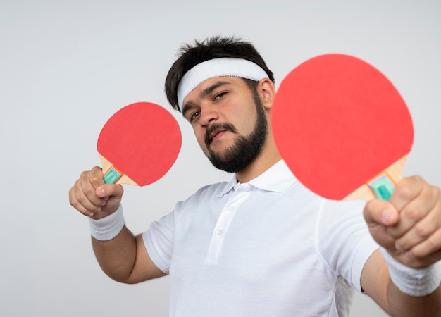 Foto gratuita hombre deportivo joven confiado con diadema y muñequera sosteniendo raquetas de ping pong aislado en la pared blanca