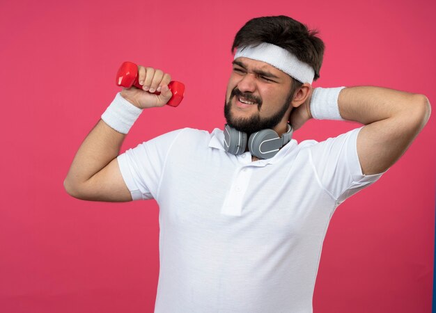 Hombre deportivo joven disgustado con diadema y muñequera con auriculares sosteniendo mancuernas y poniendo la mano en el cuello