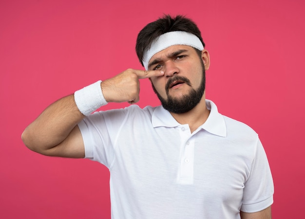 Foto gratuita hombre deportivo joven disgustado con diadema y muñequera poniendo el dedo en la nariz aislado en rosa