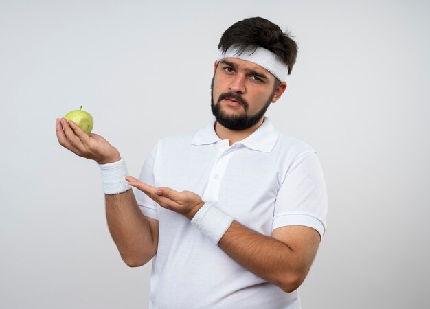 Hombre deportivo joven disgustado con diadema y muñequera sosteniendo y puntos con la mano en la manzana aislada en la pared blanca