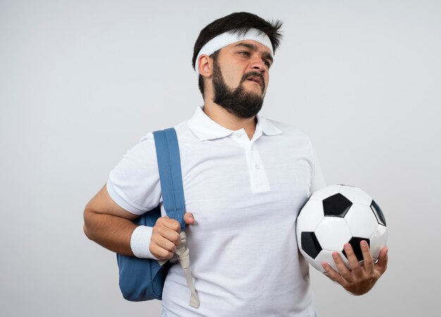 Hombre deportivo joven disgustado mirando de lado con diadema y muñequera con mochila sosteniendo la bola aislada en la pared blanca