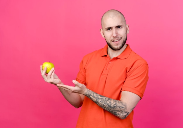 Hombre deportivo joven disgustado sosteniendo y puntos con la mano en la manzana aislada en la pared rosa
