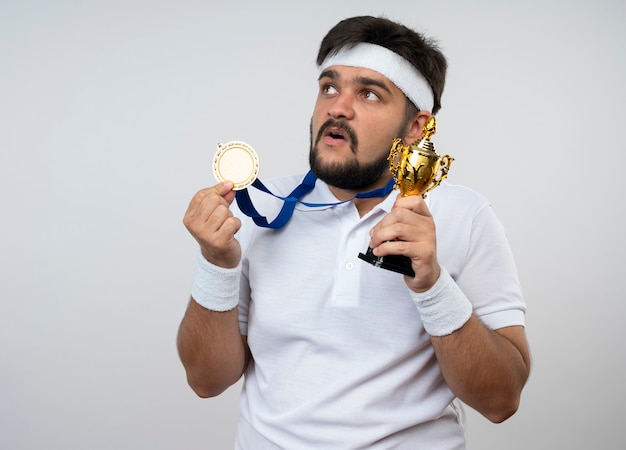 Hombre deportivo joven sorprendido con diadema y muñequera con la celebración de la copa del ganador vistiendo y sosteniendo la medalla aislada en la pared blanca