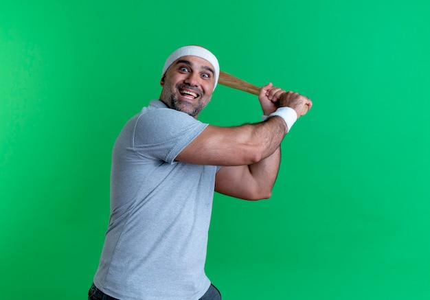 Hombre deportivo maduro en diadema balanceando un bate de béisbol sonriendo alegremente de pie sobre la pared verde