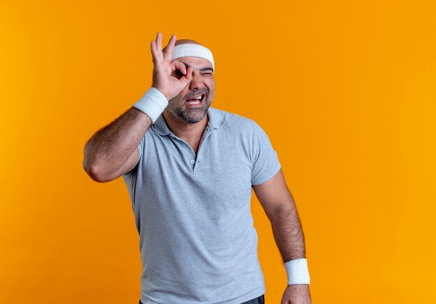 Hombre deportivo maduro en diadema haciendo bien firmar con los dedos mirando a través de este cartel de pie sobre la pared naranja