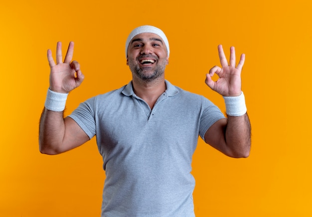 Hombre deportivo maduro en diadema mirando hacia el frente mostrando el signo de ok con ambas manos de pie sobre la pared naranja