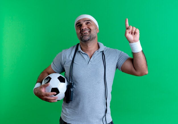 Hombre deportivo maduro en diadema sosteniendo un balón de fútbol apuntando hacia arriba con el dedo teniendo una nueva idea de pie sobre la pared verde