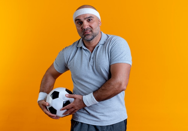 Hombre deportivo maduro en diadema sosteniendo un balón de fútbol mirando hacia el frente con cara seria de pie sobre la pared naranja