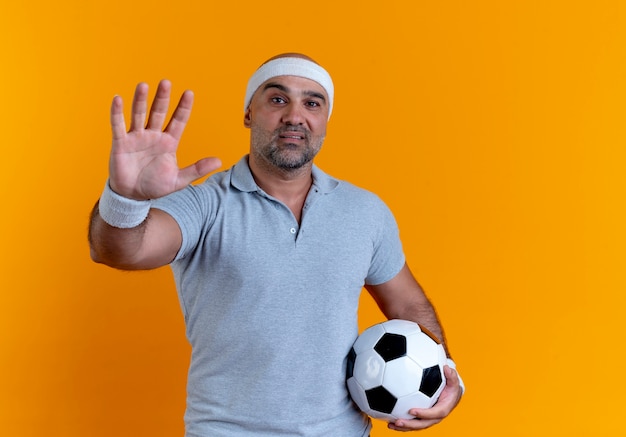 Hombre deportivo maduro en diadema sosteniendo un balón de fútbol mirando hacia el frente mostrando la palma de pie sobre la pared naranja