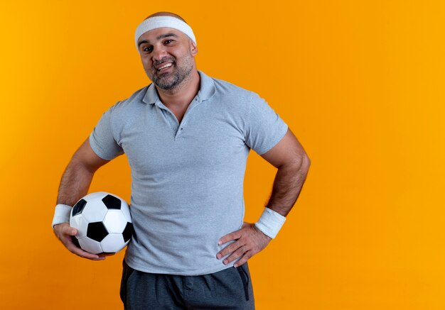 Hombre deportivo maduro en diadema sosteniendo un balón de fútbol mirando hacia el frente con una sonrisa en la cara de pie sobre la pared naranja